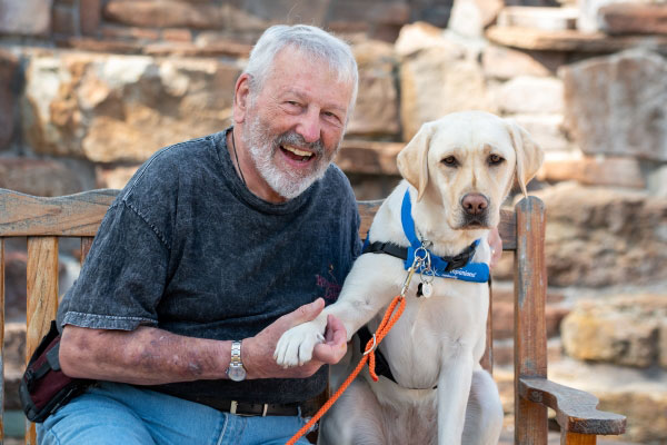 Photos shows a smiling older man seated on a bench next to a golden Labrador retriever dog wearing a service animal harness.